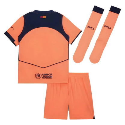 A kids' soccer kit consisting of an orange and navy blue short-sleeve jersey, matching orange shorts, and two matching orange socks with navy cuffs. The jersey features 'BARCA' and 'UNHCR ACNUR' logos.