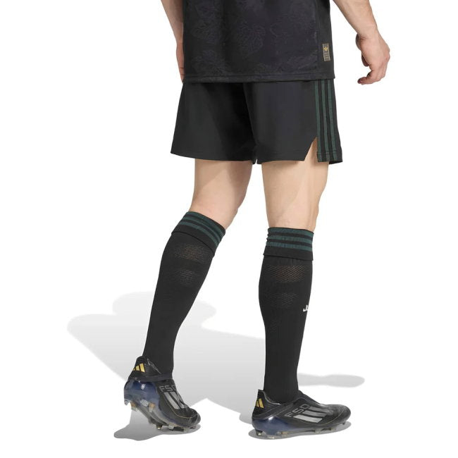 A model from the waist down wearing black soccer shorts with green stripes, black knee-high socks, and black soccer cleats on a white background.