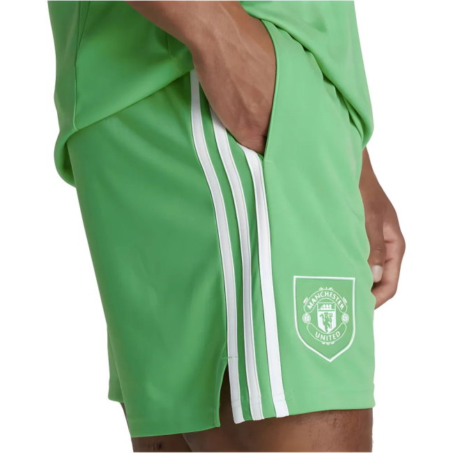 Green athletic shorts with three white side stripes and a white Manchester United crest patch, shown on a model against a white background.